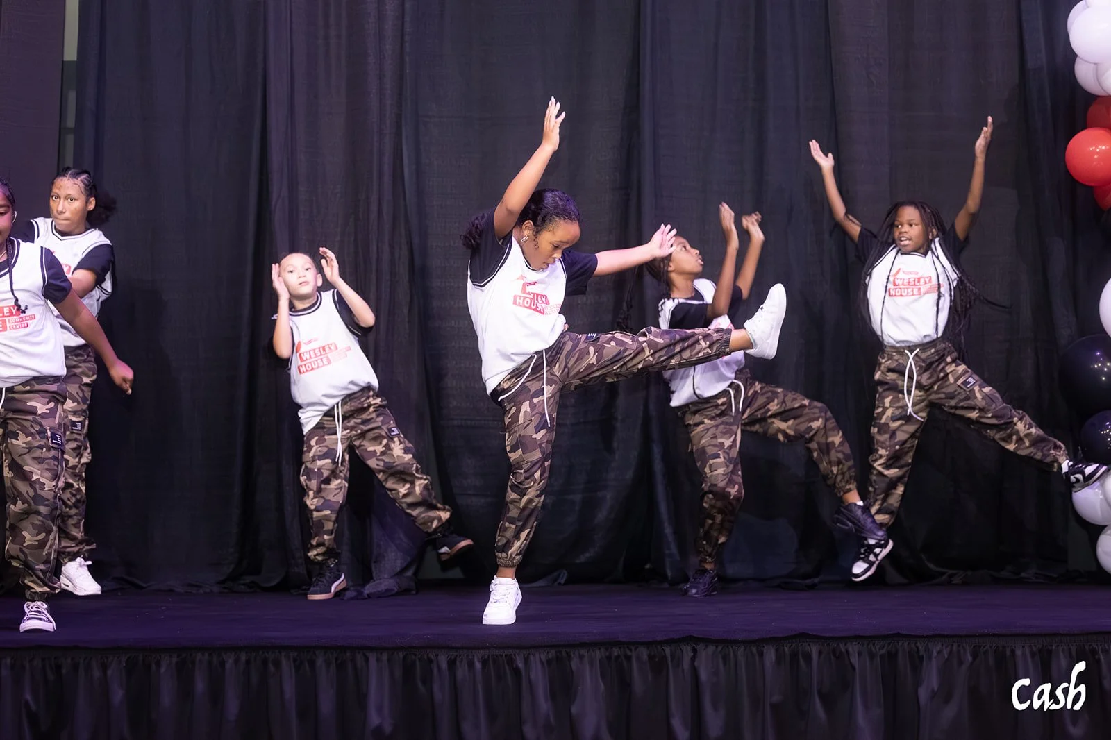 Children performing a dance on stage, wearing matching outfits with camouflage pants and white shirts with red and black text, against a black curtain backdrop, with balloons to the right.