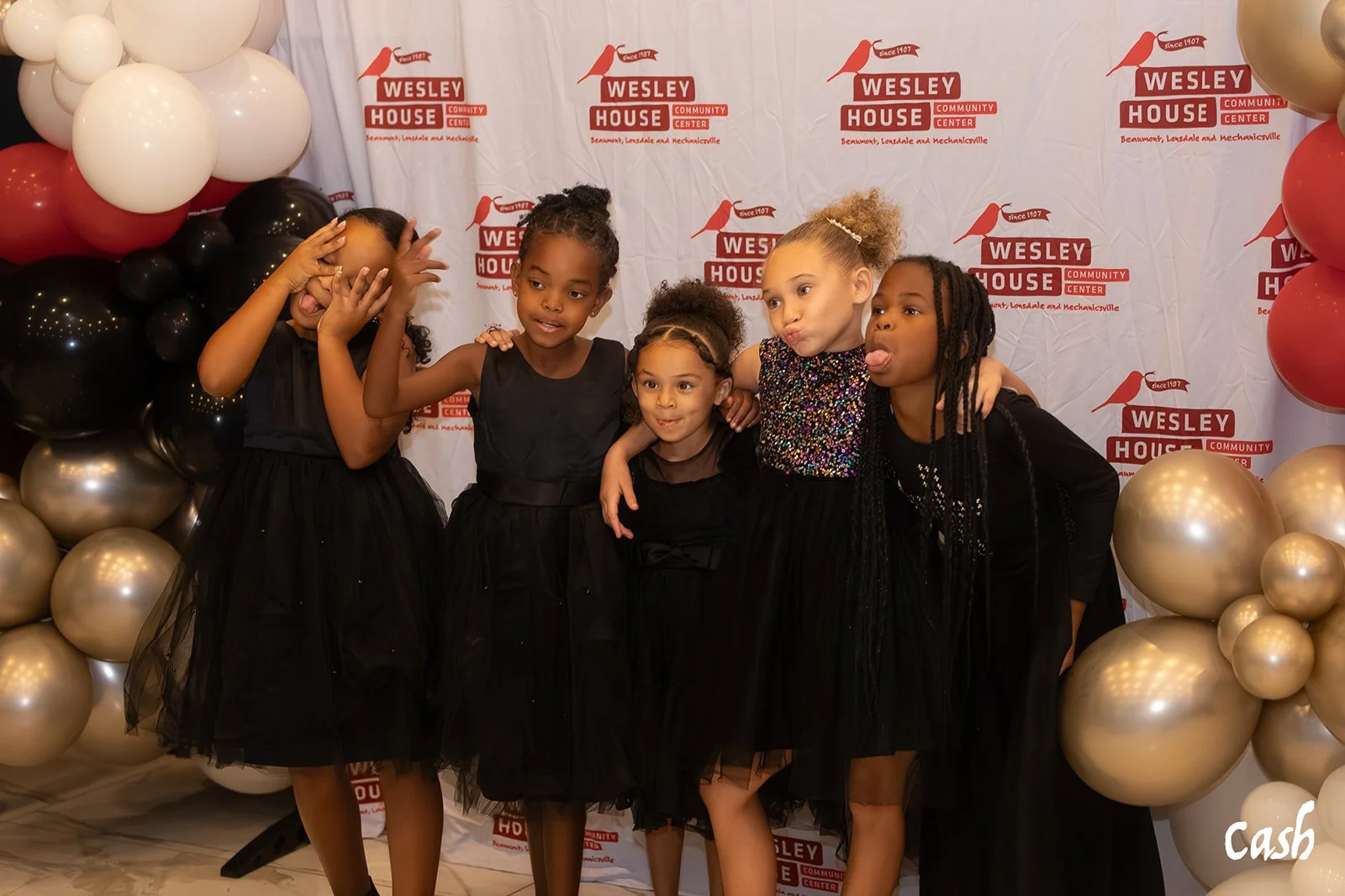 Five young girls dressed in black dresses posing together at an event with a backdrop featuring the Wesley House Community Center logo, surrounded by gold, black, white, and red balloons.