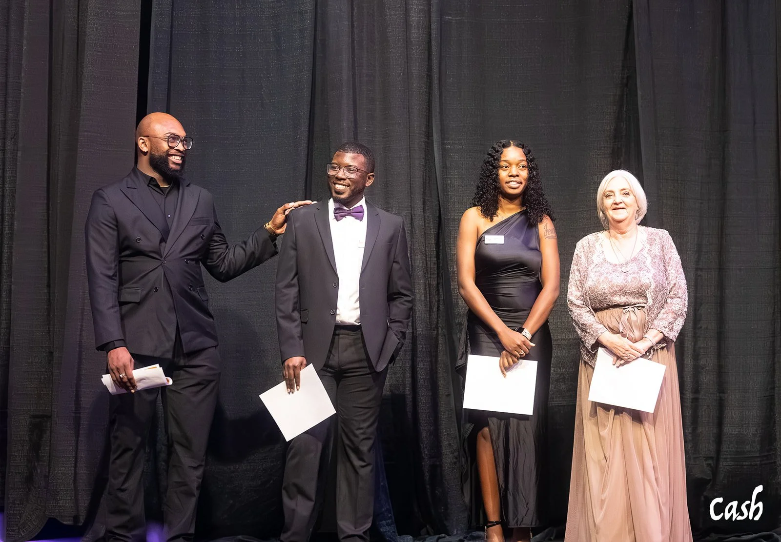 Four people standing on a stage with black curtains, dressed in formal attire. Two men and two women, holding white papers. One man is touching the shoulder of the other, all smiling, at what appears to be an award ceremony or celebration.