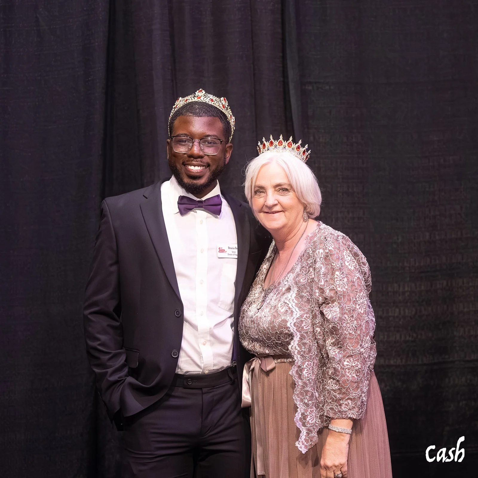 A young man and an older woman standing together, both wearing crowns, at a formal event against a black backdrop.