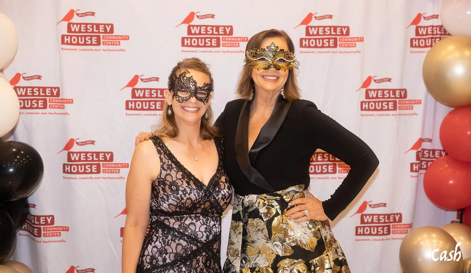 Two women in dresses and masks posing in front of a Wesley House Community Center backdrop with gold and black balloons on both sides.
