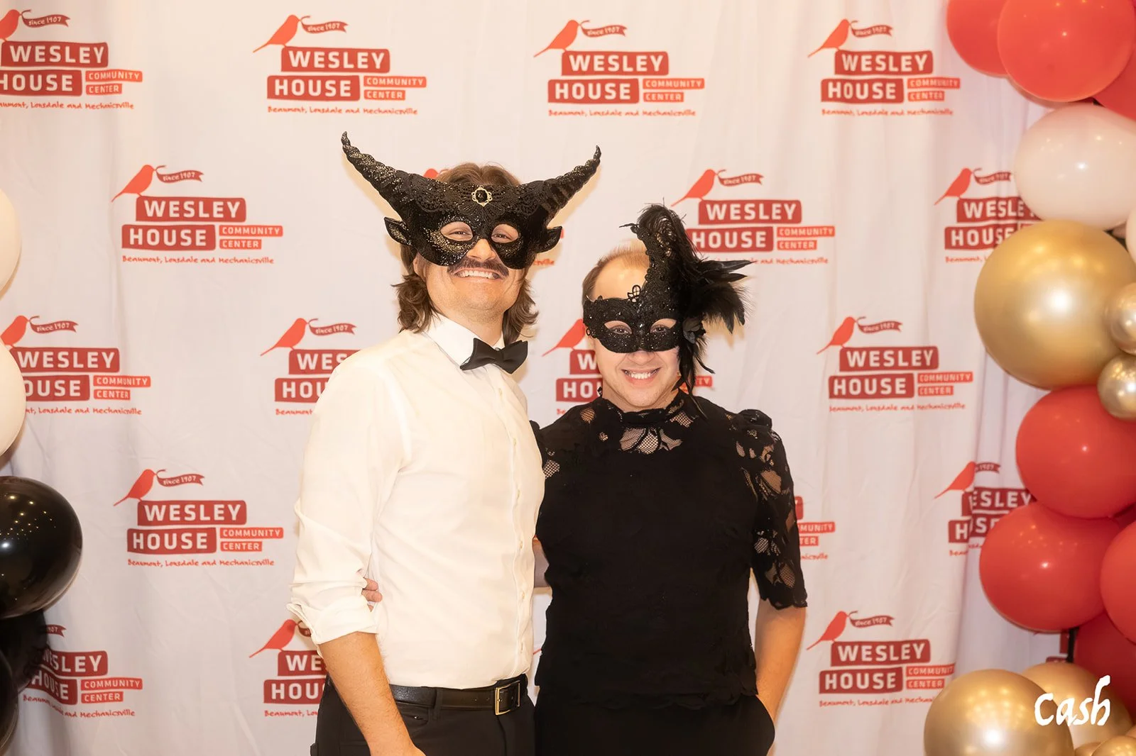 Two people dressed in formal attire wearing black masquerade masks smiling in front of a Wesley House Community Center backdrop with balloons on the side.