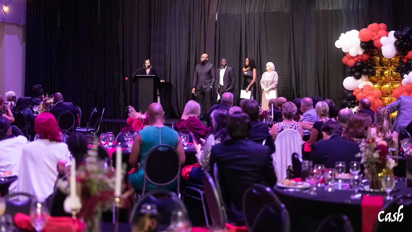 An awards ceremony on a stage with five people standing and a woman speaking at a podium, while an audience watches. The stage is decorated with balloon arrangements in black, white, and red, and there are tables with floral centerpieces in the foreg