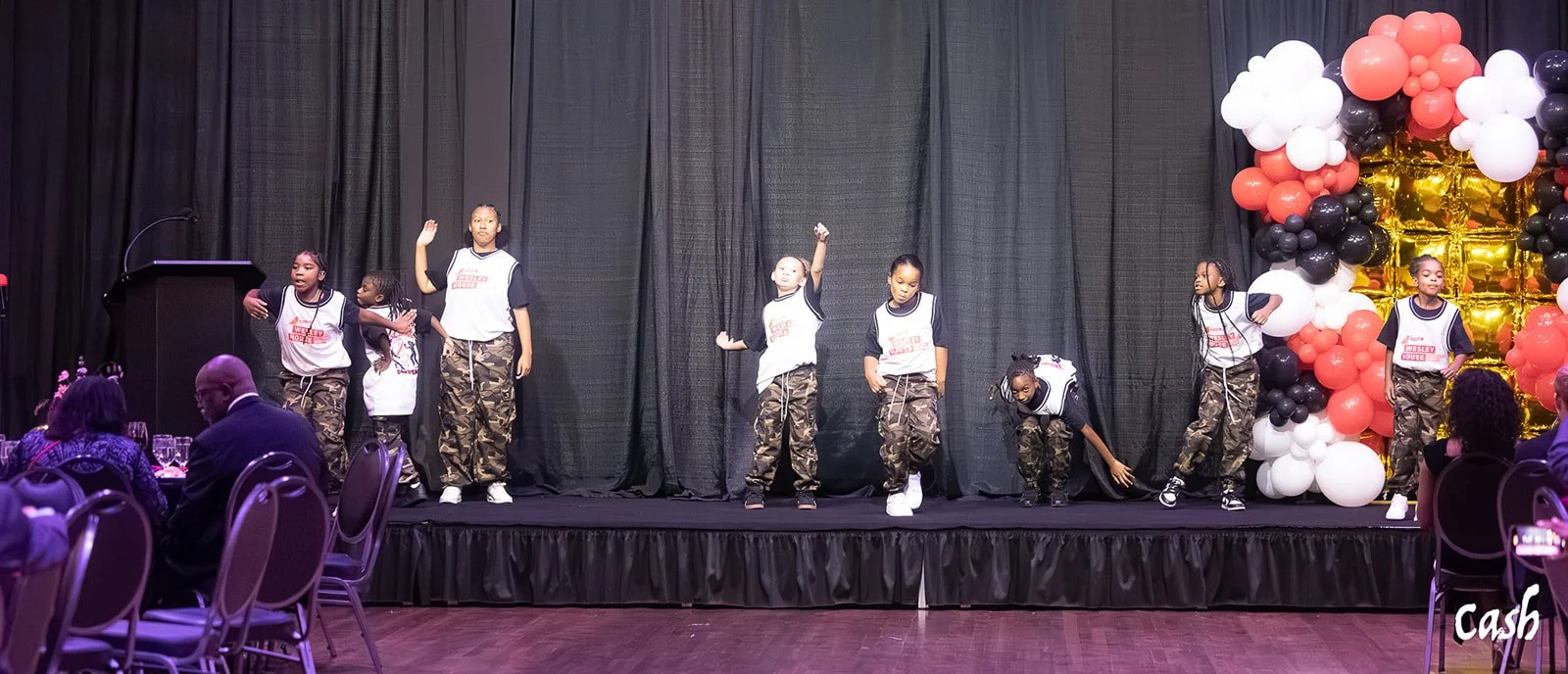 Children performing a dance on stage in front of an audience, with balloons decorating the stage area.