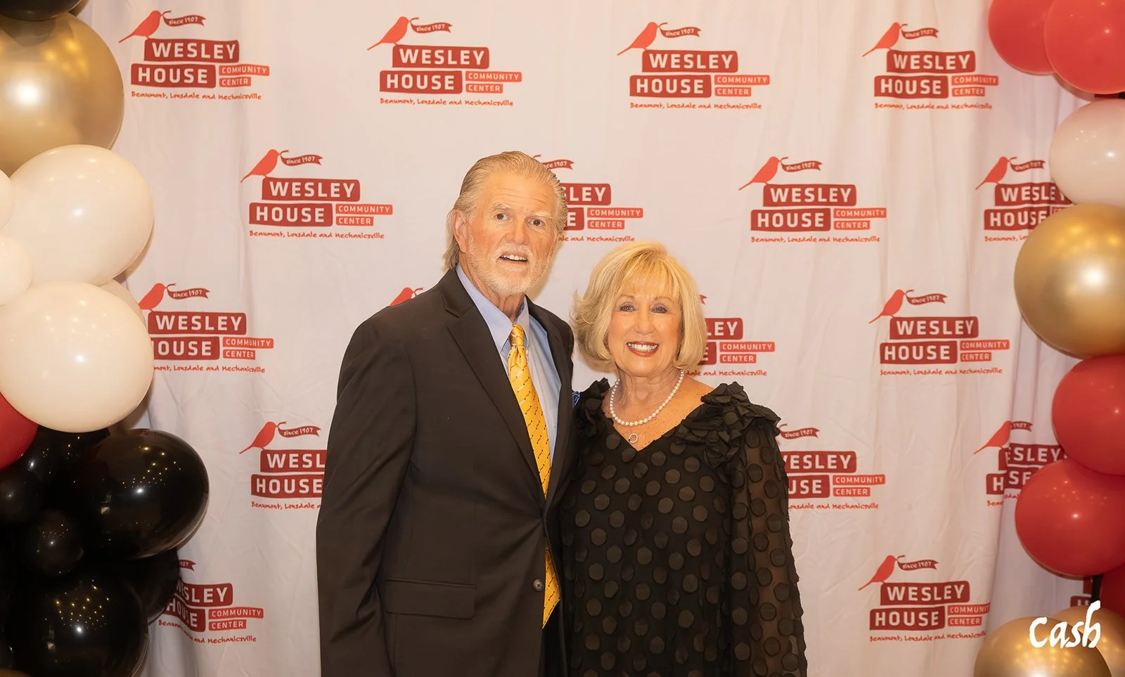 A man and a woman dressed in formal attire standing in front of a Wesely House Community Center backdrop with balloons on the sides.