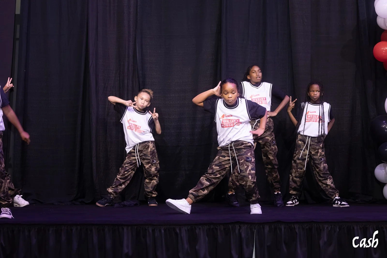 Group of young girls performing a dance on stage, wearing camouflage pants and white shirts with a logo, with black curtains and balloons in the background.