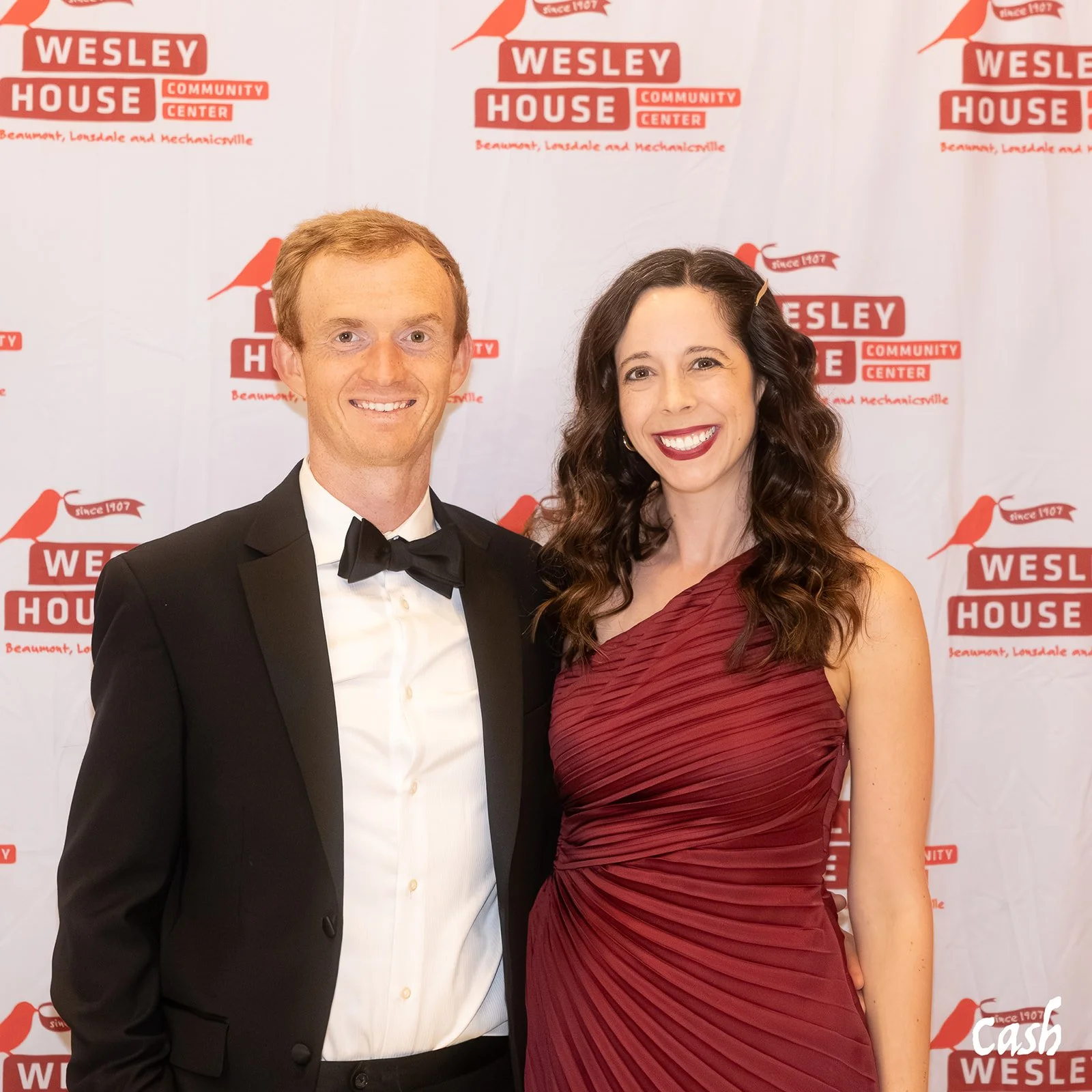 A man in a tuxedo and a woman in a red gown standing in front of a backdrop with 'WESLEY HOUSE COMMUNITY CENTER' logos, smiling for the camera.
