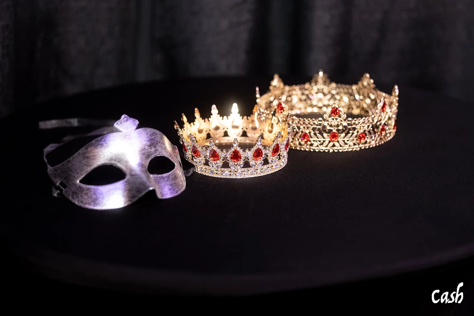 A silver masquerade mask and two ornate crowns with red gemstones and diamonds are displayed on a black surface.