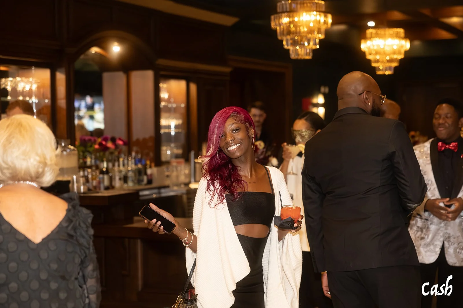 A group of diverse people socializing at an upscale bar or lounge. A woman with pink hair holding a drink and a phone smiling at the camera. Others are dressed elegantly, and the setting features warm lighting and chandeliers.