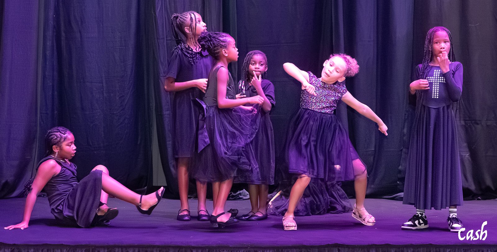 Seven young girls on stage dressed in black, performing a dance or play with a black curtain backdrop.