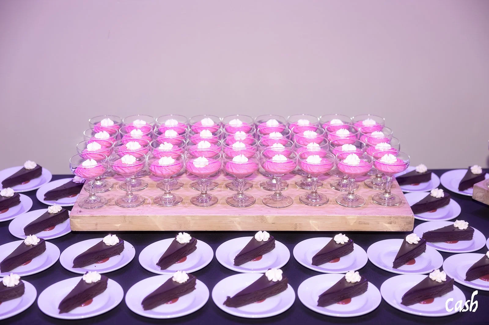 Dessert table with pink parfaits in coupe glasses on a wooden board and chocolate cake slices on white plates arranged around the table