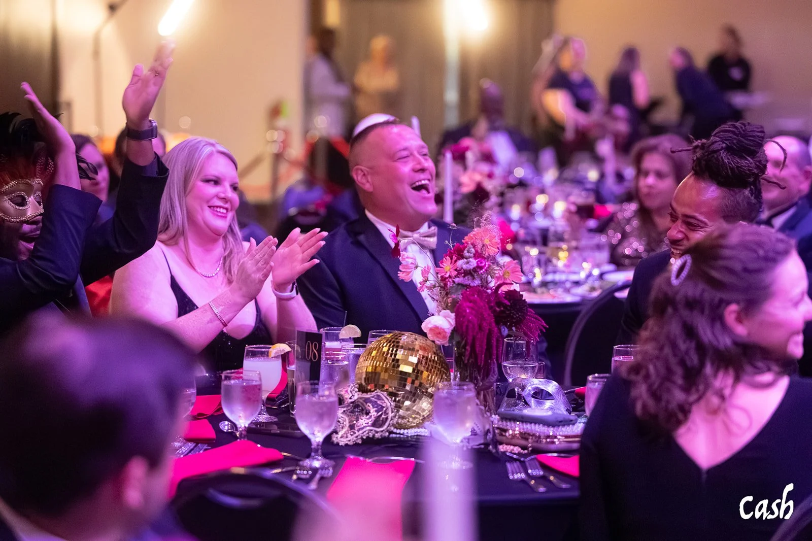 People celebrating at a formal event with seated guests clapping and smiling. The table features floral arrangements, gold disco ball, glasses of water, and decorative masks. The atmosphere is festive with purple lighting.
