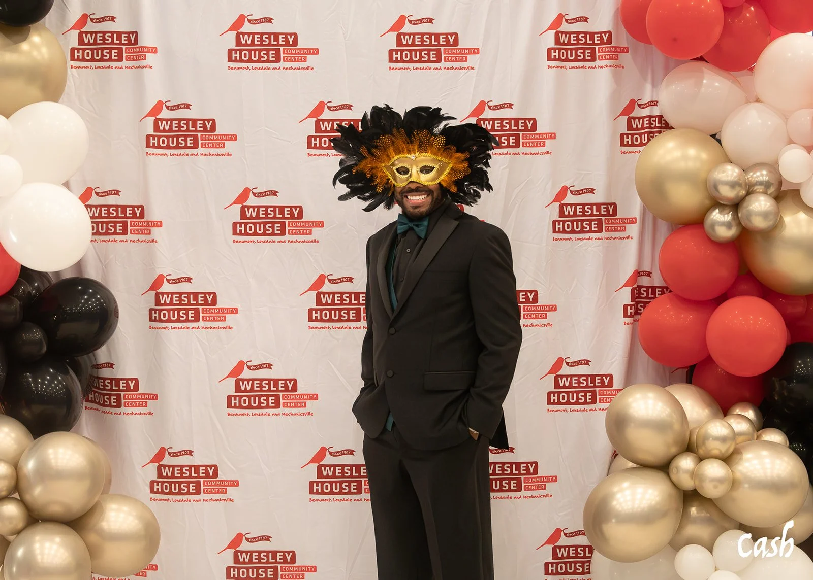 A man in a black suit and teal shirt wearing a gold and black Mardi Gras mask, standing in front of a backdrop with Wesley House logos, with party balloons on each side.