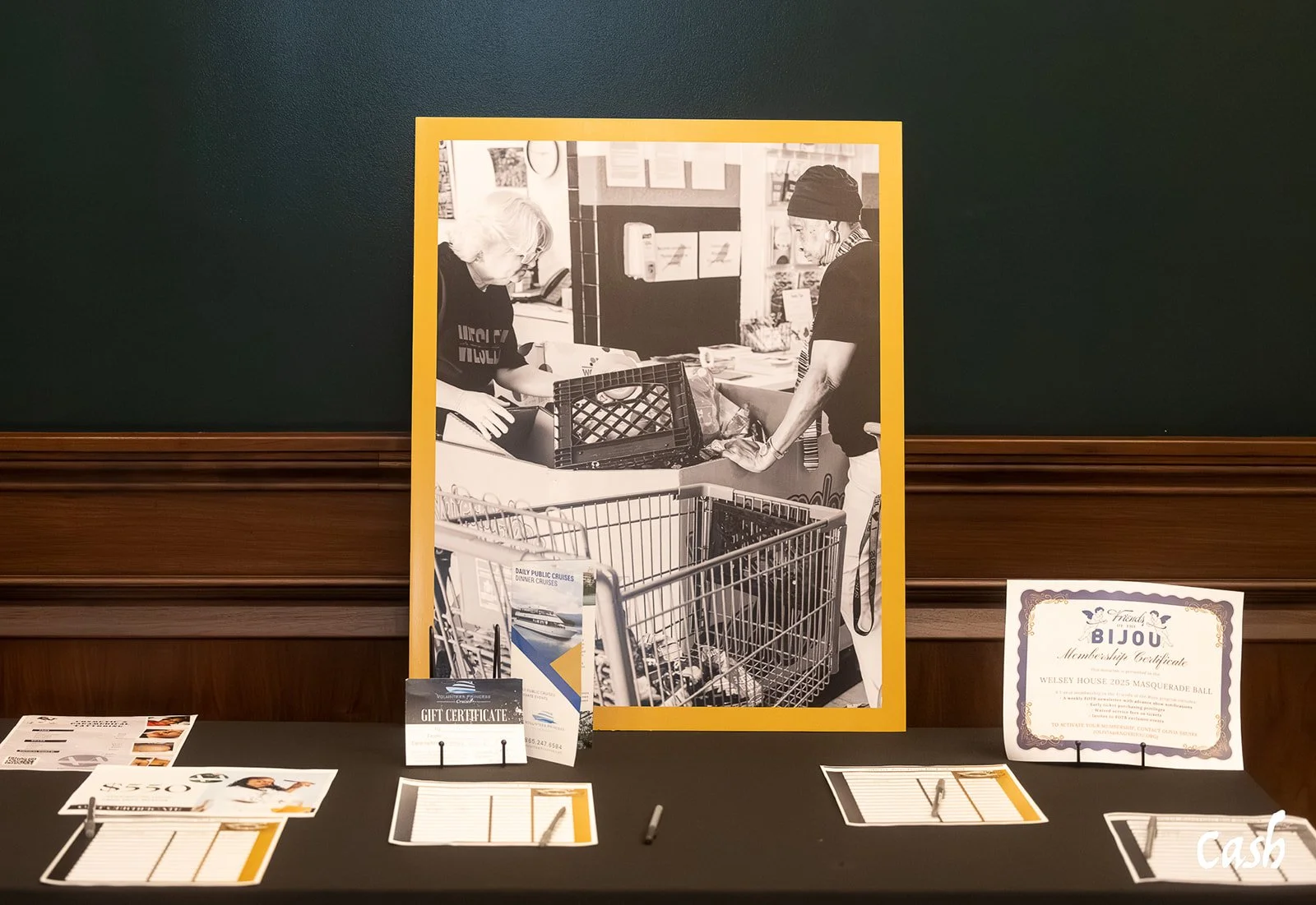 A black-and-white photo of two people working in a grocery store or market stall, with shopping carts and various items on display. The photo is framed with a yellow border and is part of a display table that includes certificates, flyers, and sign-i