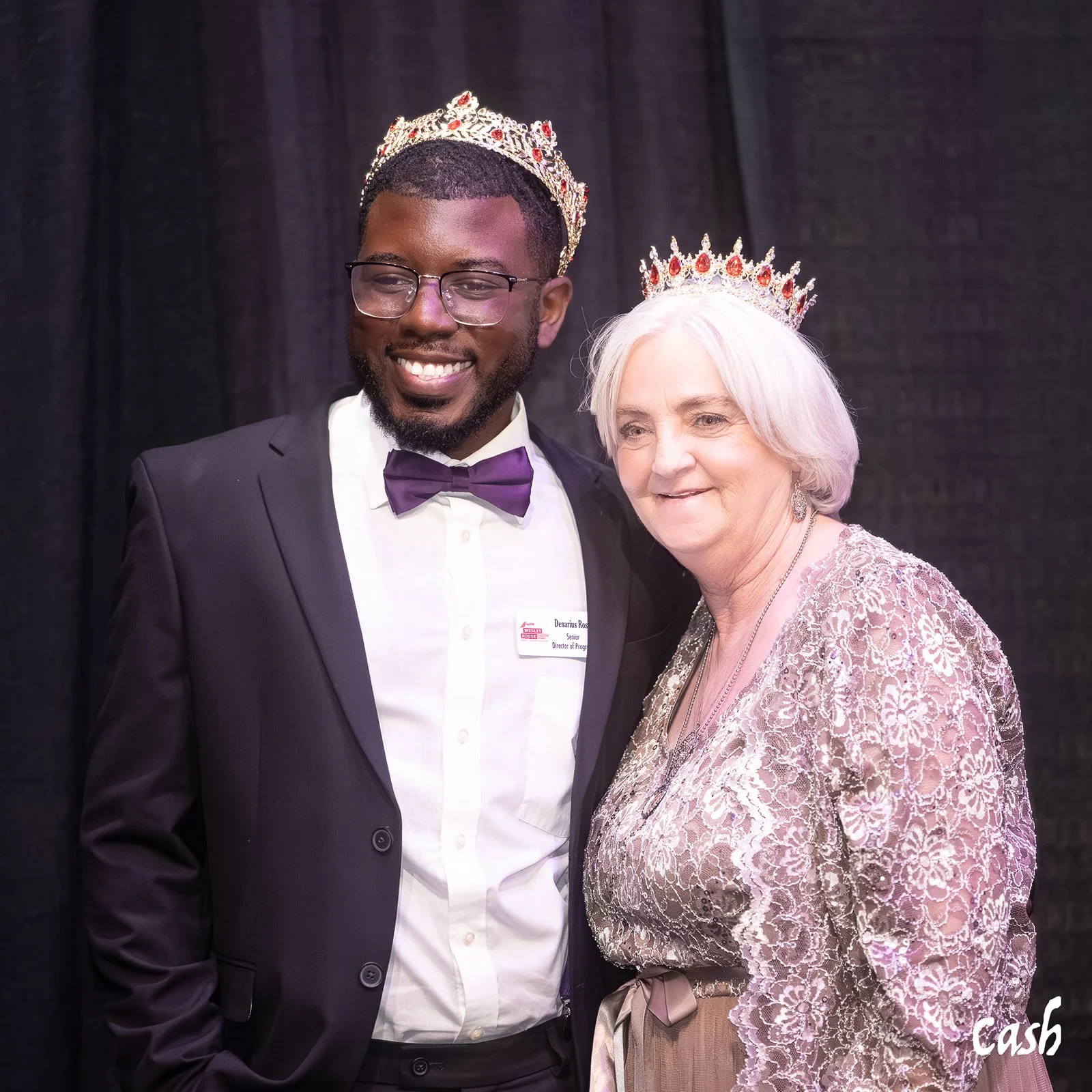A young man and an older woman wearing crowns and formal attire standing together at a celebration or event, smiling.