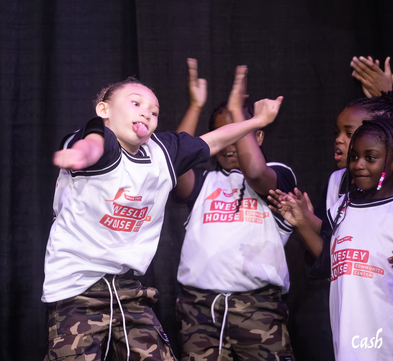 Children wearing Wesley House Community Center T-shirts and camouflage pants clapping and dancing on stage.