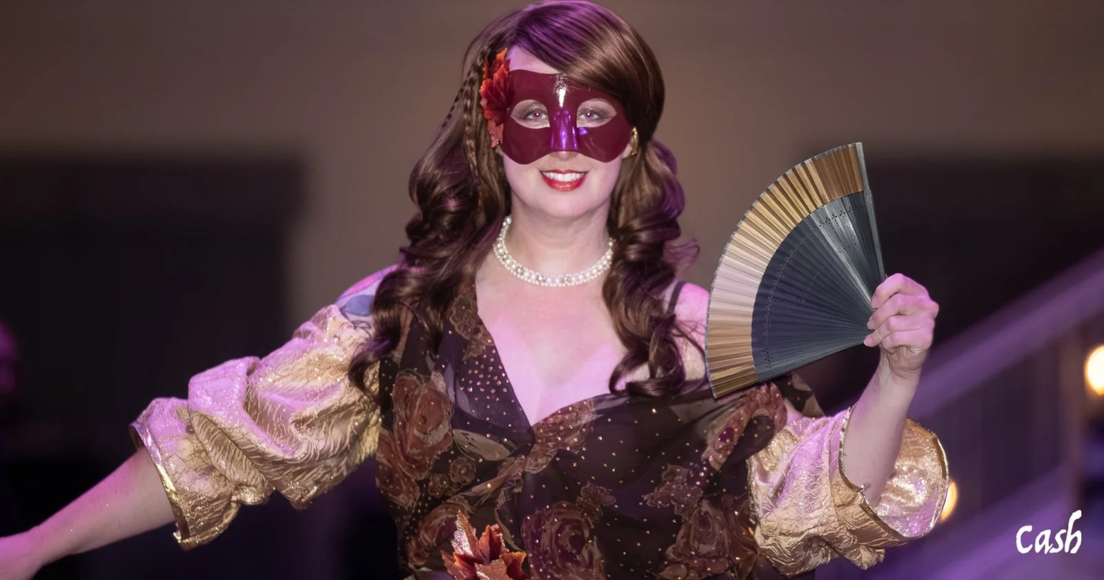 A woman dressed in a colorful, floral-patterned dress with gold accents, wearing a red mask and a pearl necklace, holding a folding fan.