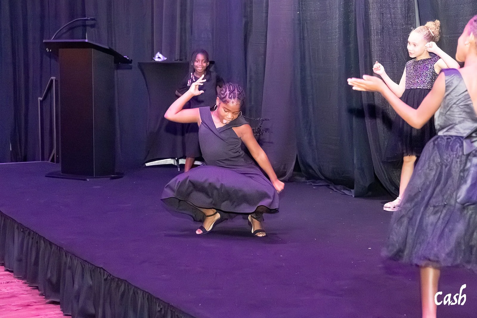 Young girls performing on a stage, dancing and smiling, with black curtains in the background.