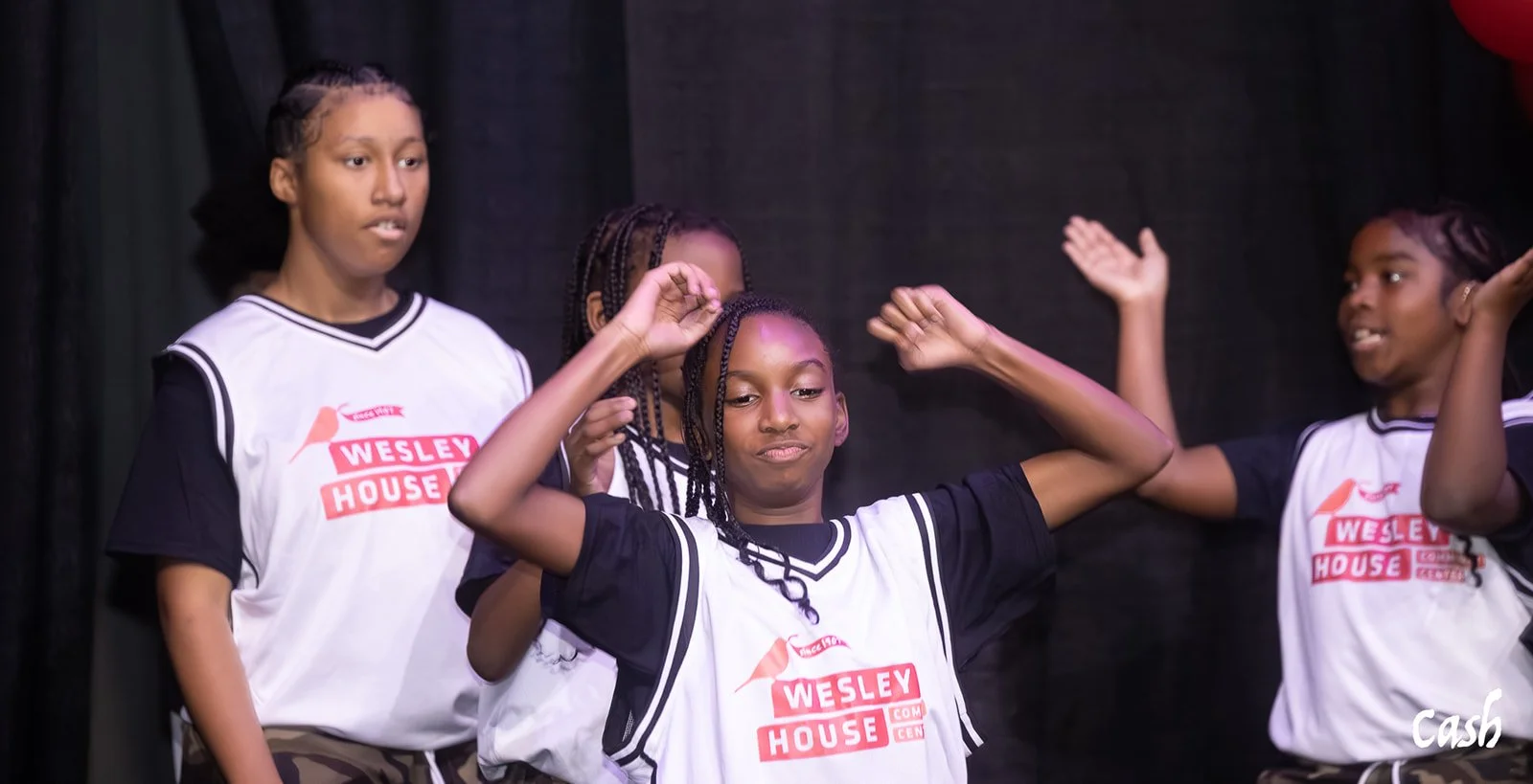 Group of young girls wearing Wesley House basketball jerseys, some with braided hair, standing on stage with dark background, some with hands raised and one with arms flexed.