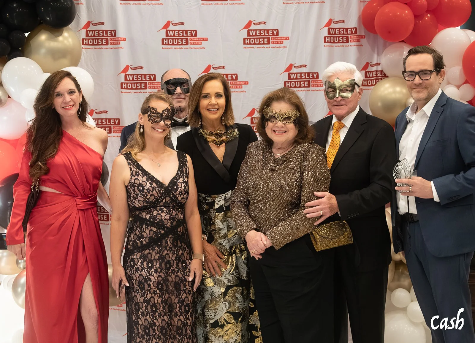 Group of eight people dressed in formal attire at a festive event with a backdrop reading 'Wesley House Community Center' and balloon decorations, some wearing masks.