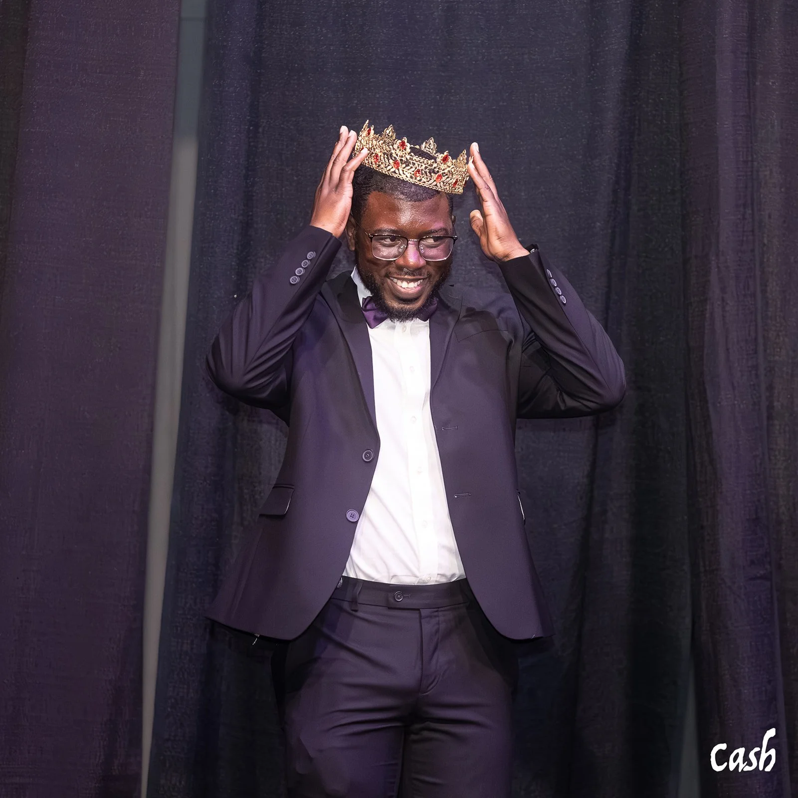 A man in a black suit and glasses is smiling while putting a decorative crown on his head during an event.