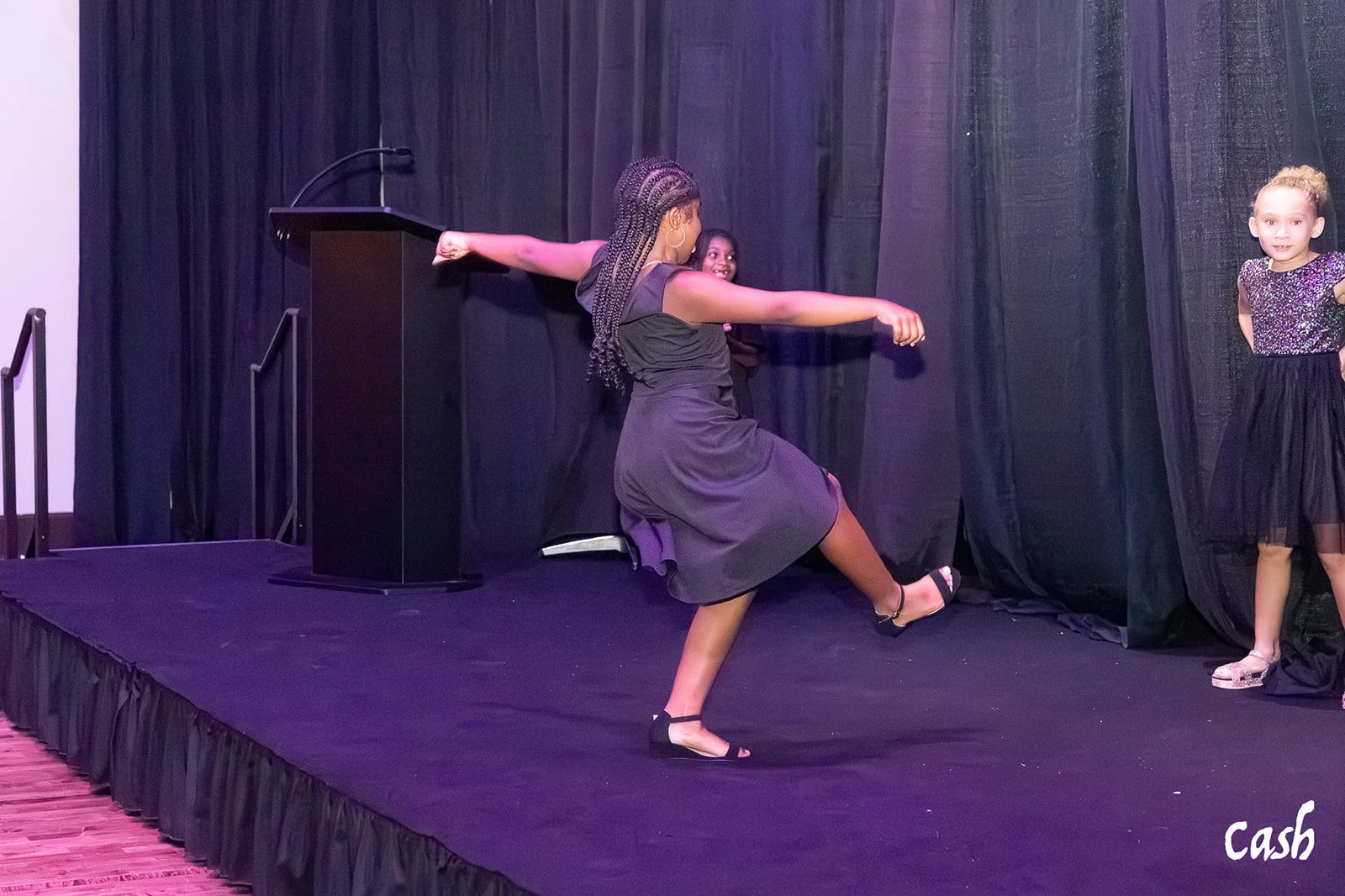 A woman on stage performing a dance move with one leg lifted and arms extended, while two young girls watch nearby. The stage has a black curtain backdrop and a podium is visible. The word 'cash' is written in the bottom right corner.