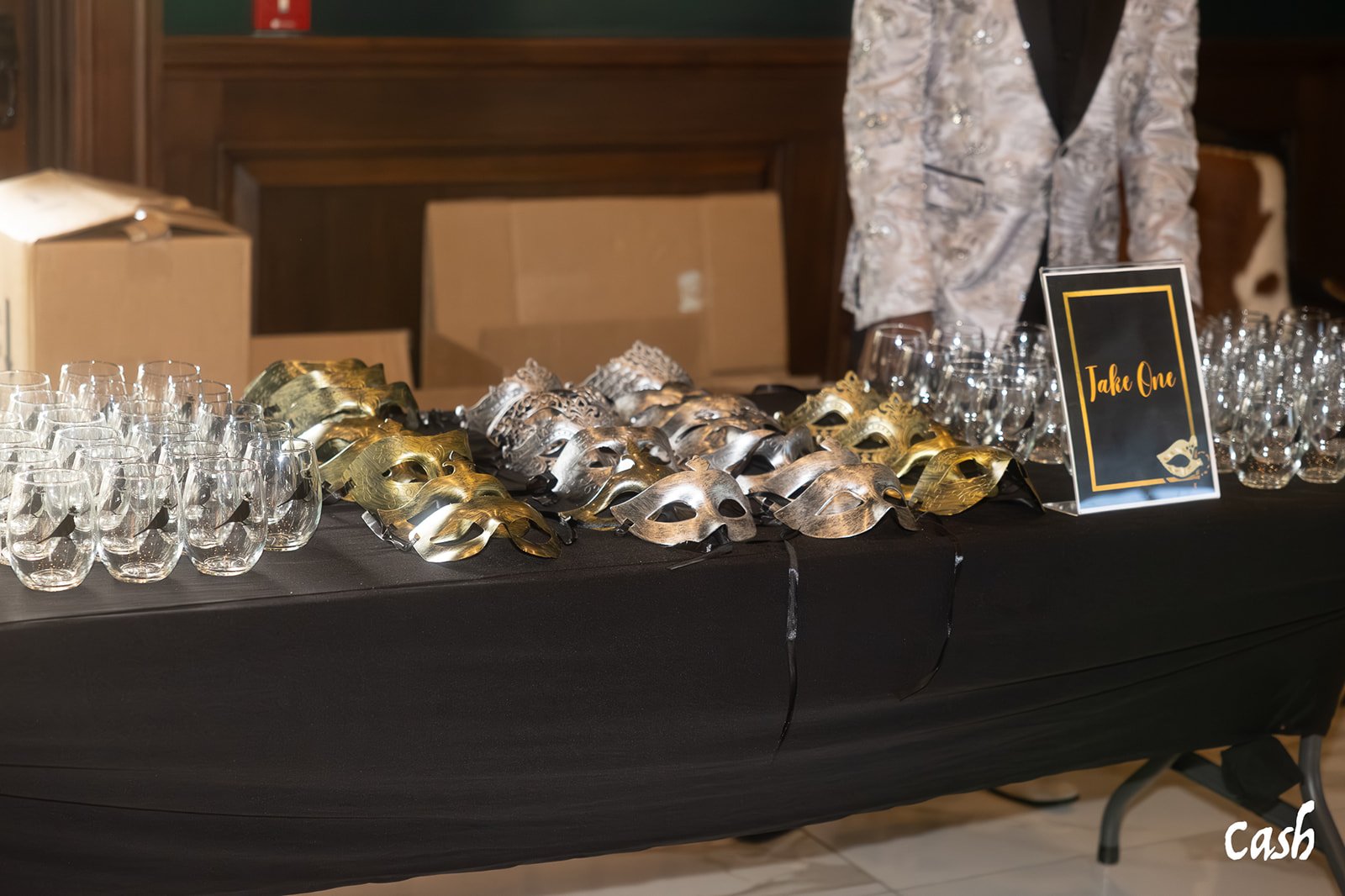 Table with gold and silver masquerade masks, wine glasses, and a black sign that reads 'Take One' at a masquerade event.