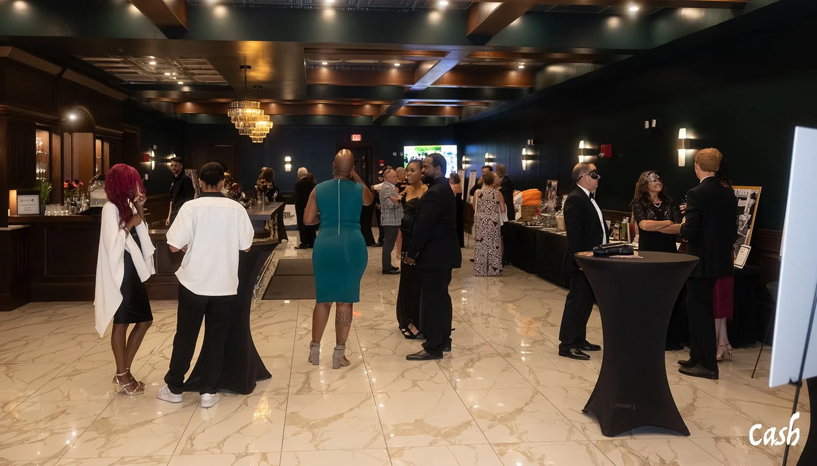 Guests socializing in a dark, elegant event space with high ceilings, chandeliers, and a marble floor. Some people are standing around tall cocktail tables, wearing formal and semi-formal attire.