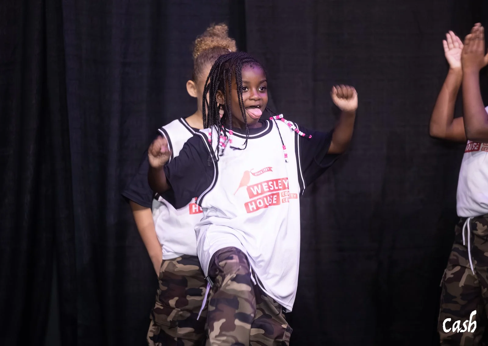Young girl with braided hair and camouflage pants dancing on stage, sticking out her tongue, wearing a Velcro sports jersey, with other children dancing beside her, against a black curtain background.