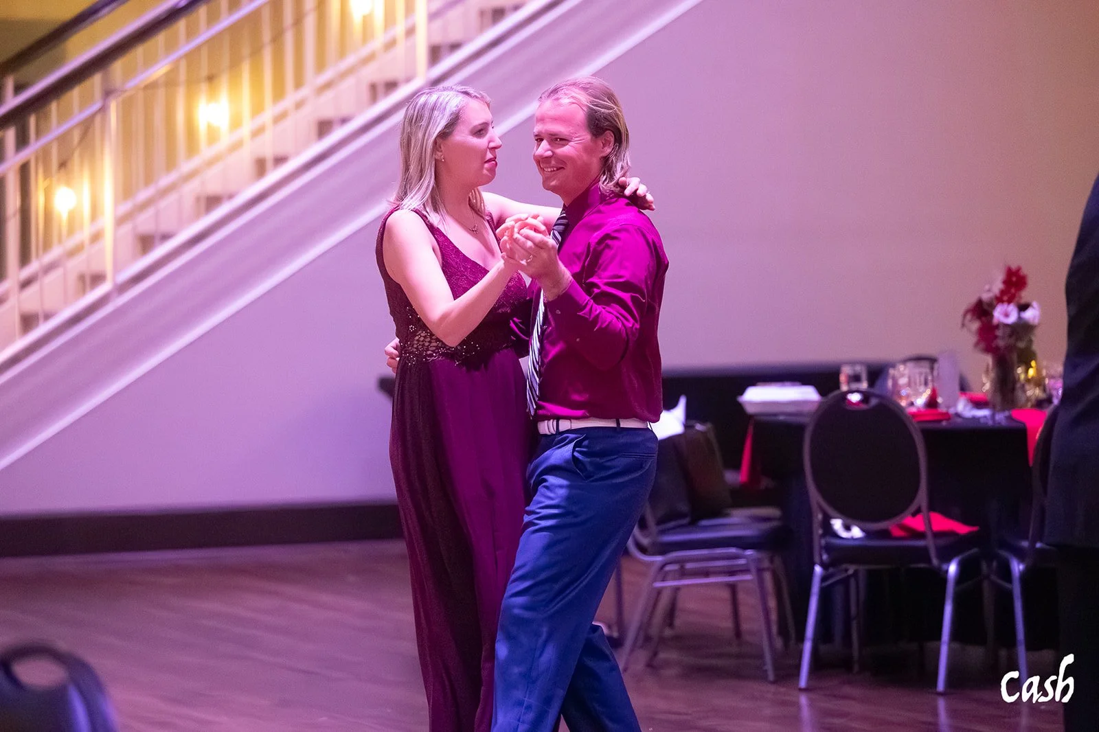 A couple dancing together at a formal event, with the woman in a purple gown and the man in a purple shirt and blue pants. The background features a staircase with yellow railings and decorated tables.