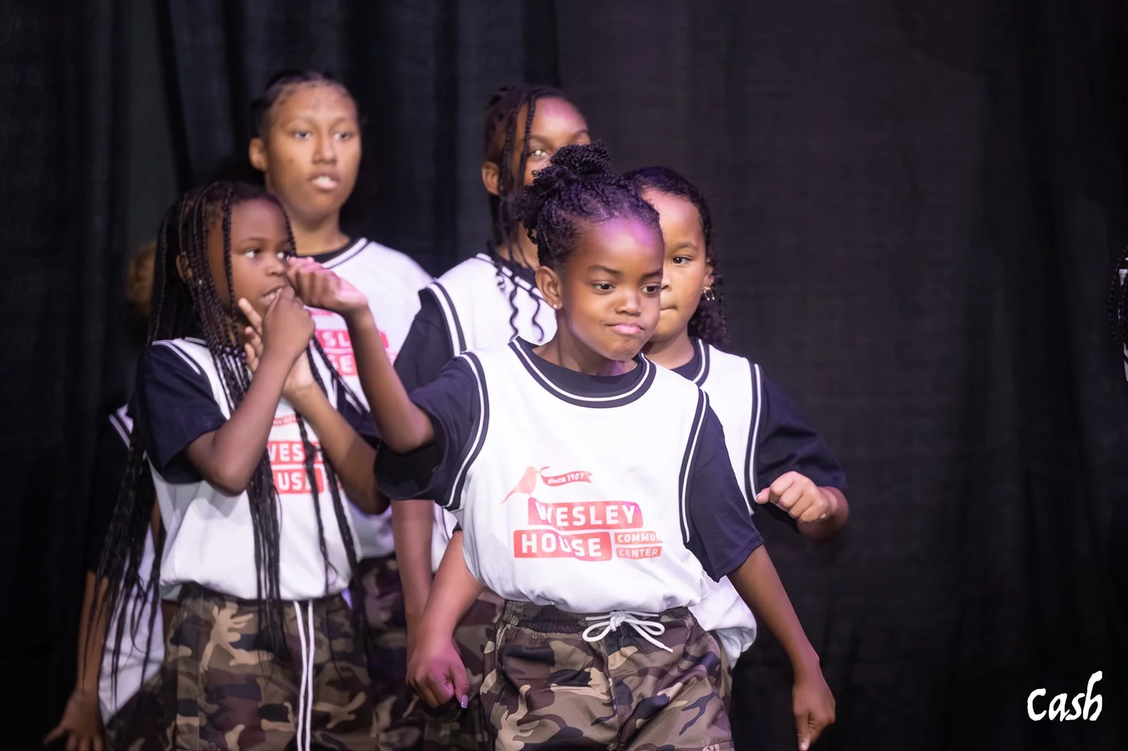 Group of young children performing on stage, some with fists raised, wearing black and white shirts with 'Wesley House' logo, and camouflage pants, against a dark background.