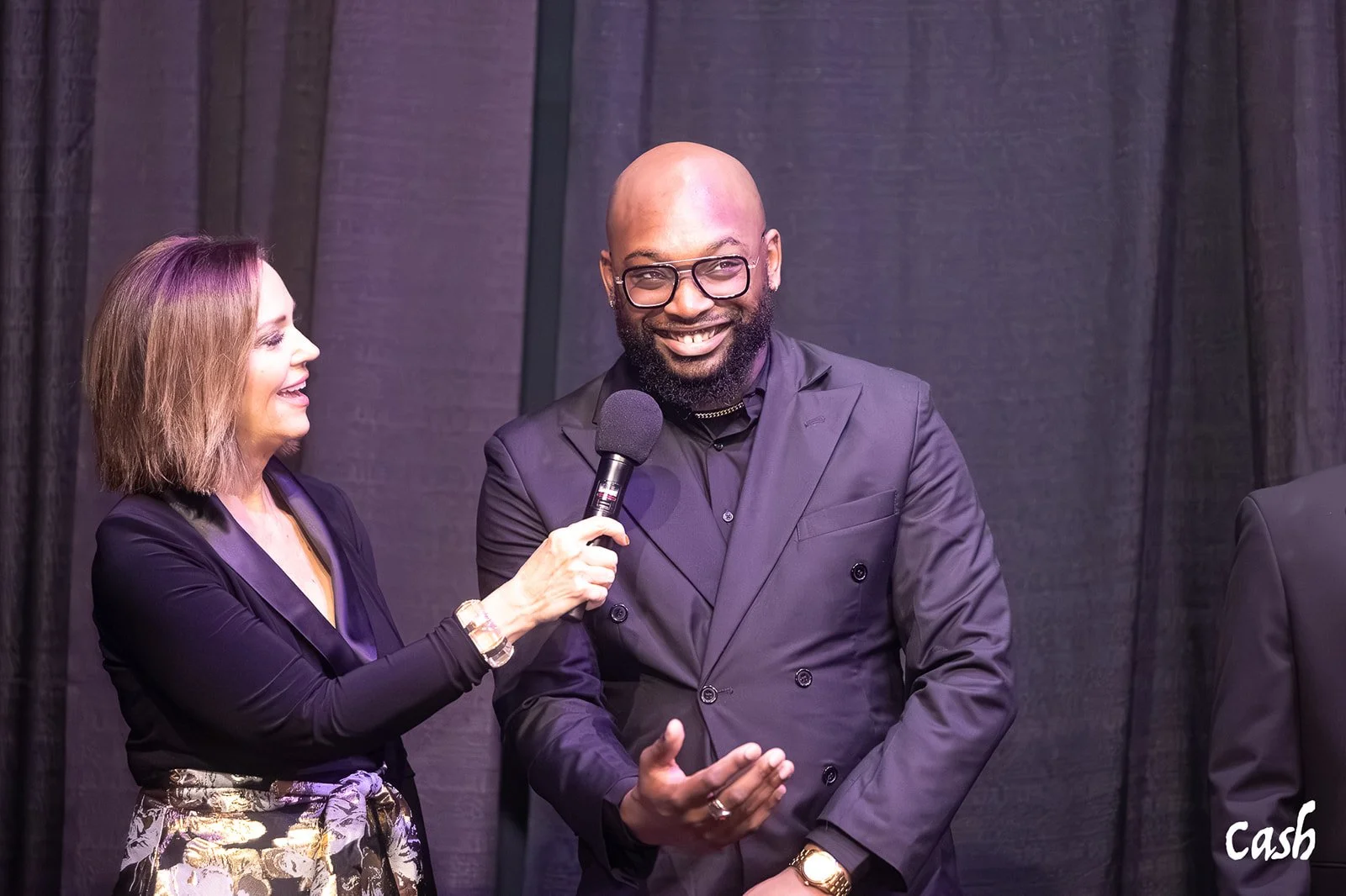 A woman in black clothing with a patterned skirt holding a microphone and smiling while interviewing a man in a dark suit, glasses, and a gold watch at an event.