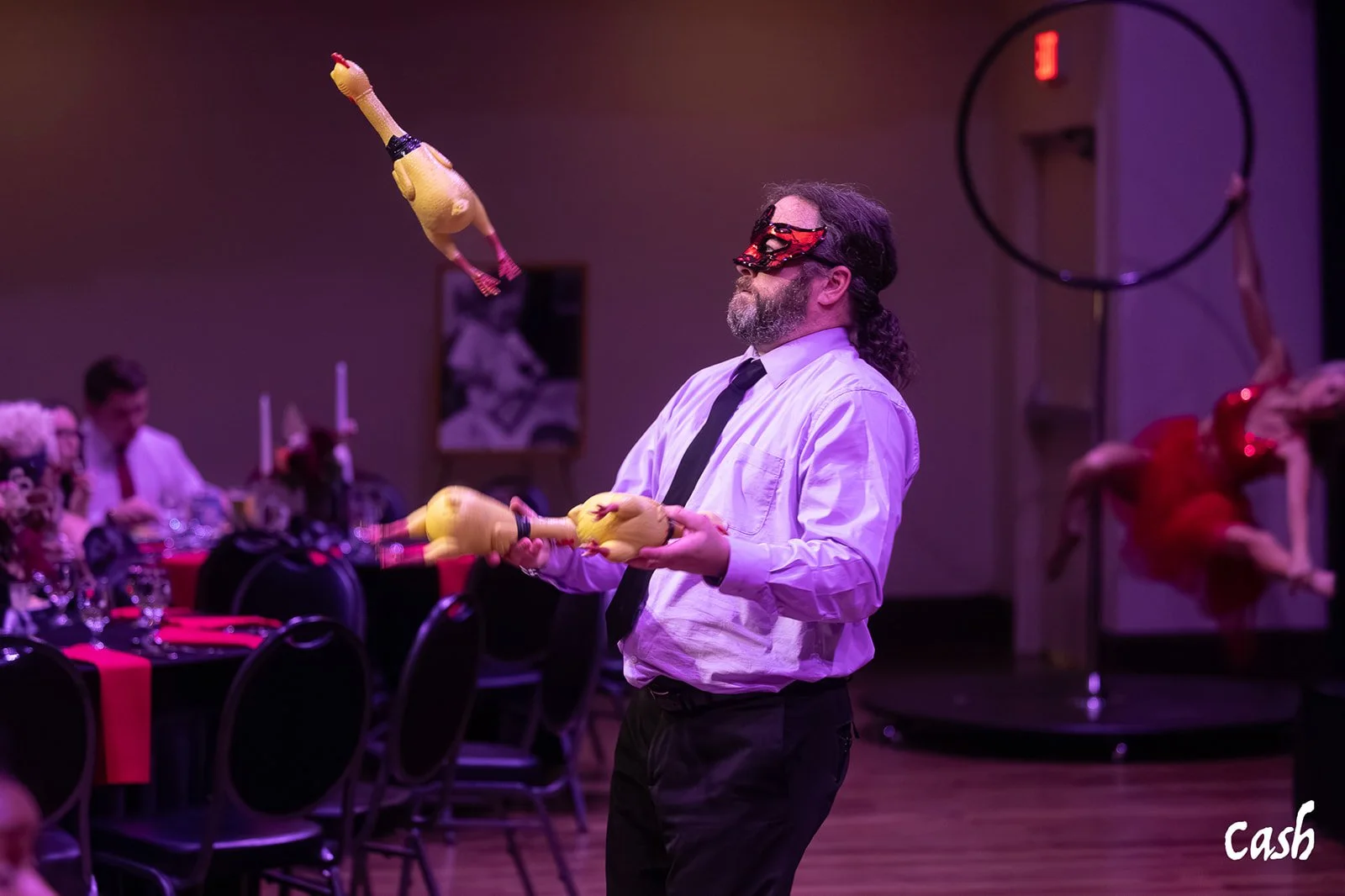 A man with long hair and a beard, wearing a white shirt, black tie, and decorative red mask, balances yellow rubber chickens on his hands during a performance in a dimly lit room decorated for a dinner event.