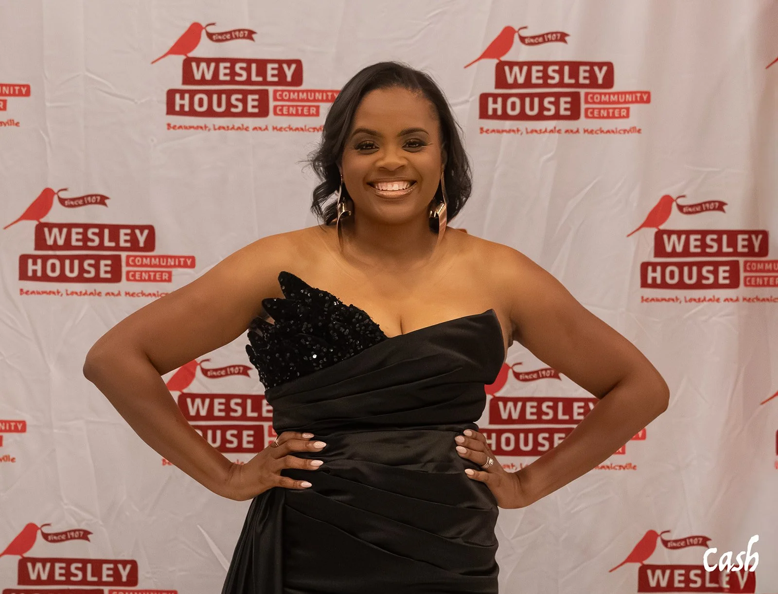 A woman in a black strapless dress with a sequined ruffle on the shoulder, smiling, standing in front of a Wesley House Community Center backdrop.