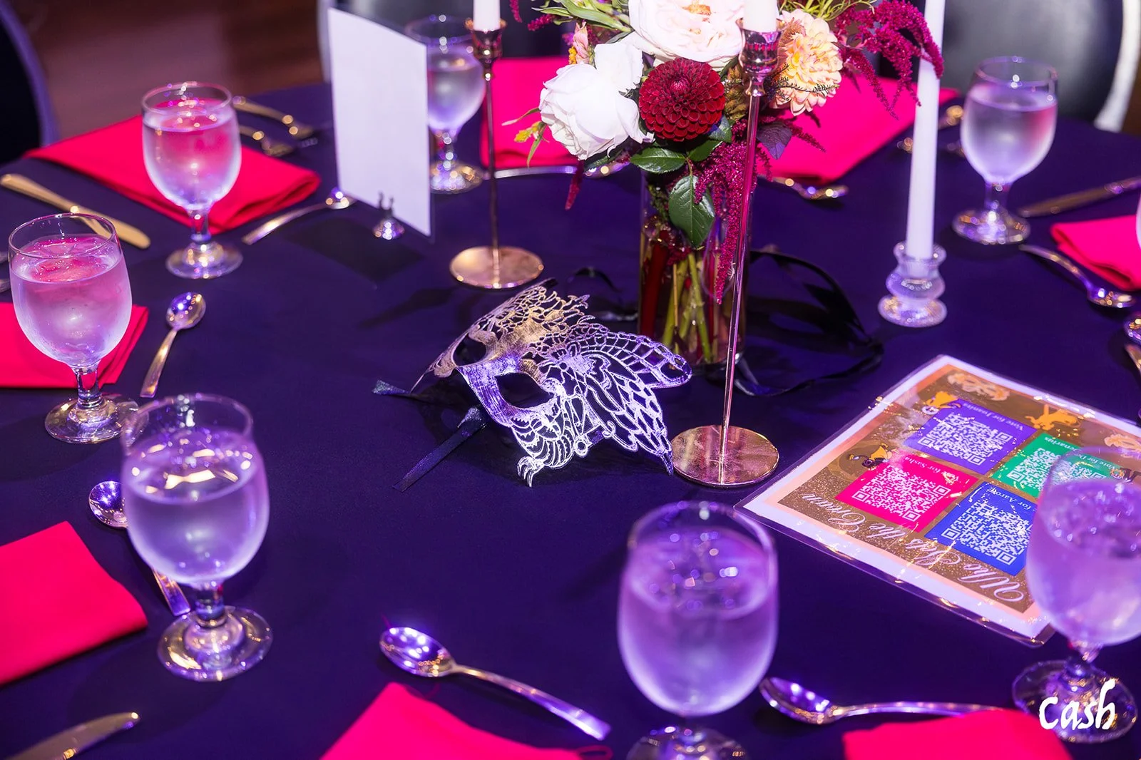 A round table set for a formal event with black tablecloth, pink and red napkins, water glasses, silverware, a floral centerpiece with pink, white, and red flowers, and a decorative metallic butterfly mask.