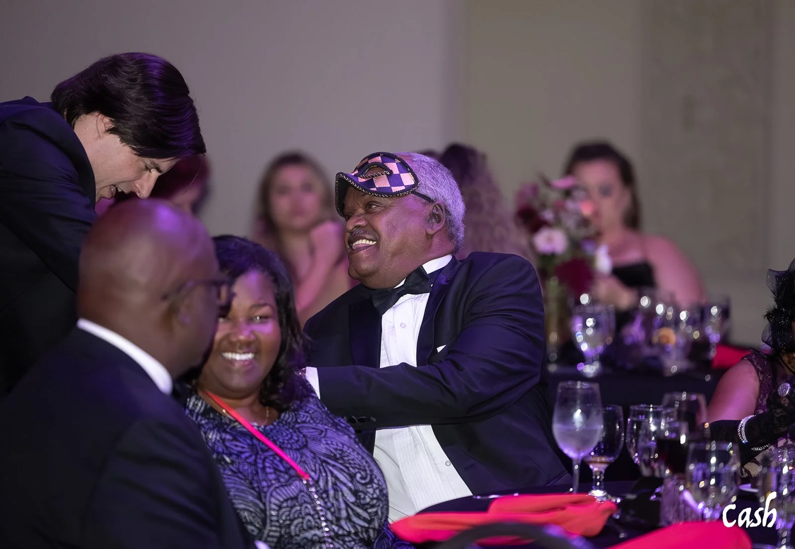 People dressed in formal tuxedos and evening gowns gathered around a banquet table at a social event, engaging in conversation and laughter.