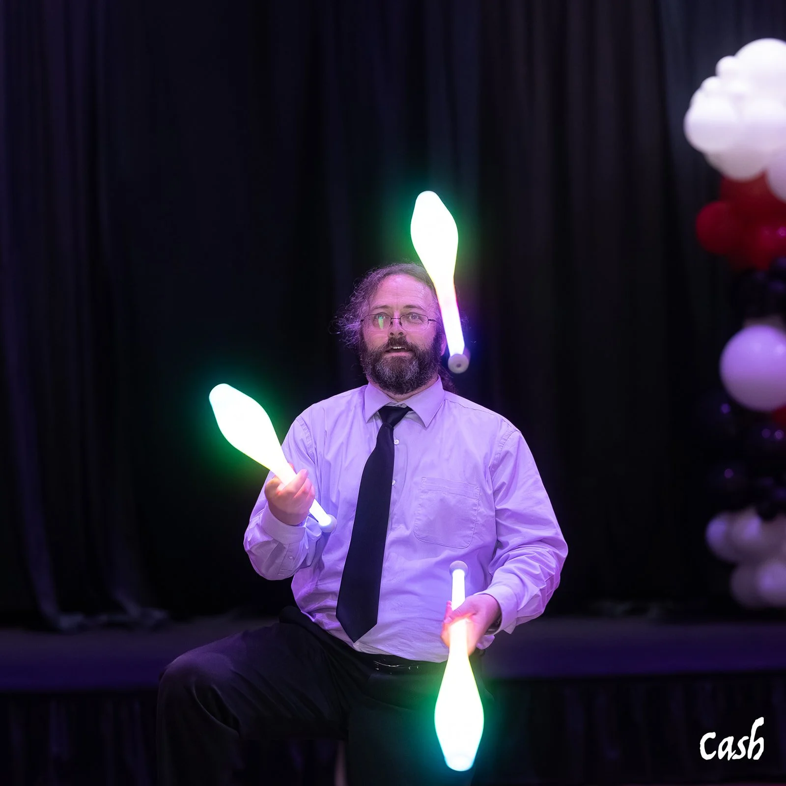 A man with glasses and a beard, wearing a dress shirt and tie, juggles glowing clubs with green, purple, and yellow lights on a stage with balloons in the background.