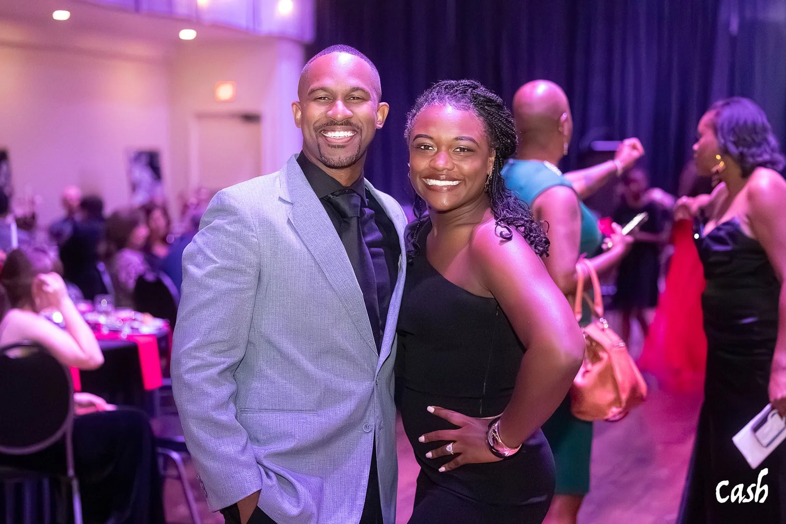 A smiling man in a gray suit and a woman in a black dress pose together at an indoor social event with many other people in the background.