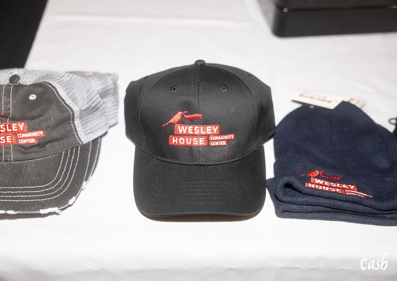 Three Wesley House Community Center hats and a folded navy beanie on a table, with a white tablecloth.
