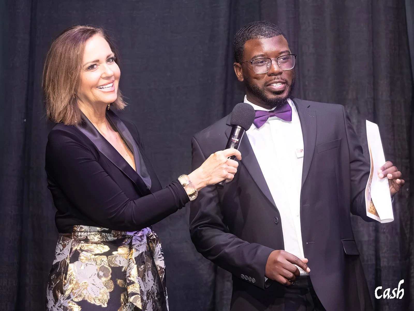 A woman with shoulder-length brown hair smiling, holding a microphone towards a man in a tuxedo with glasses, a purple bow tie, and holding papers, against a black backdrop.