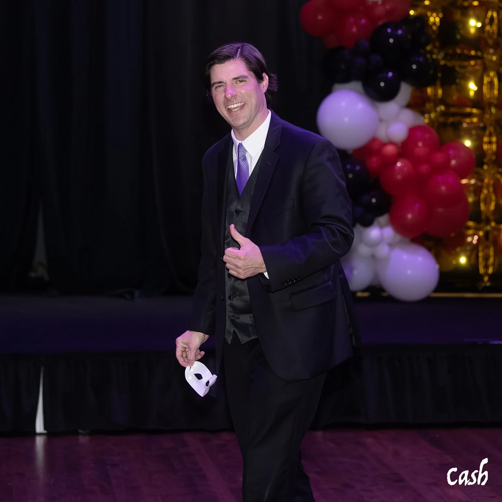 A man in a black suit with a purple tie and vest, holding a white mask in his left hand, standing on a stage with black curtains and a balloon decoration in black, red, white, and gold in the background, smiling and giving a thumbs-up.