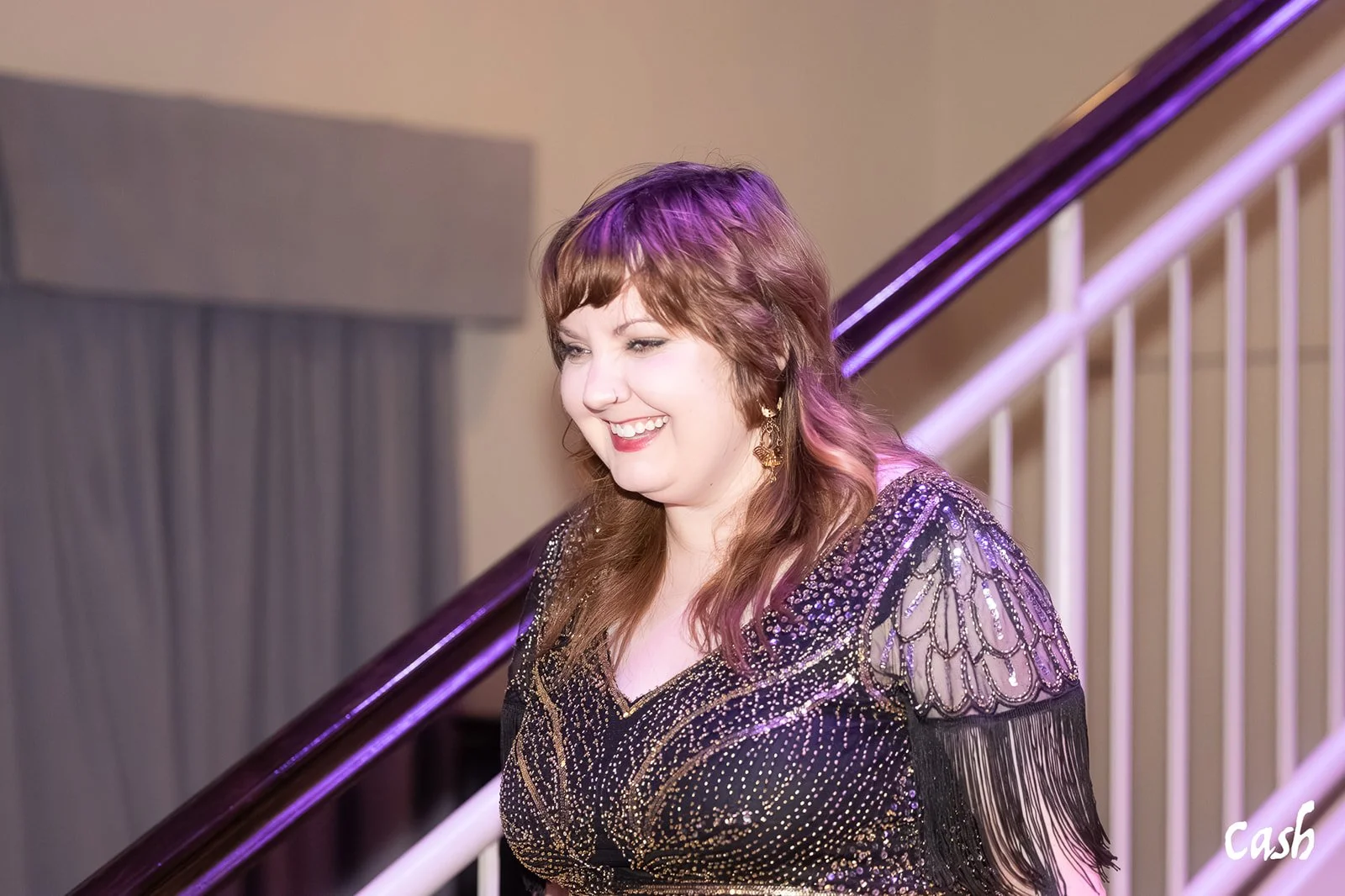 A woman with wavy, shoulder-length hair, smiling, wearing a sequined black dress with sheer, embroidered sleeves, standing near a staircase with purple lighting.