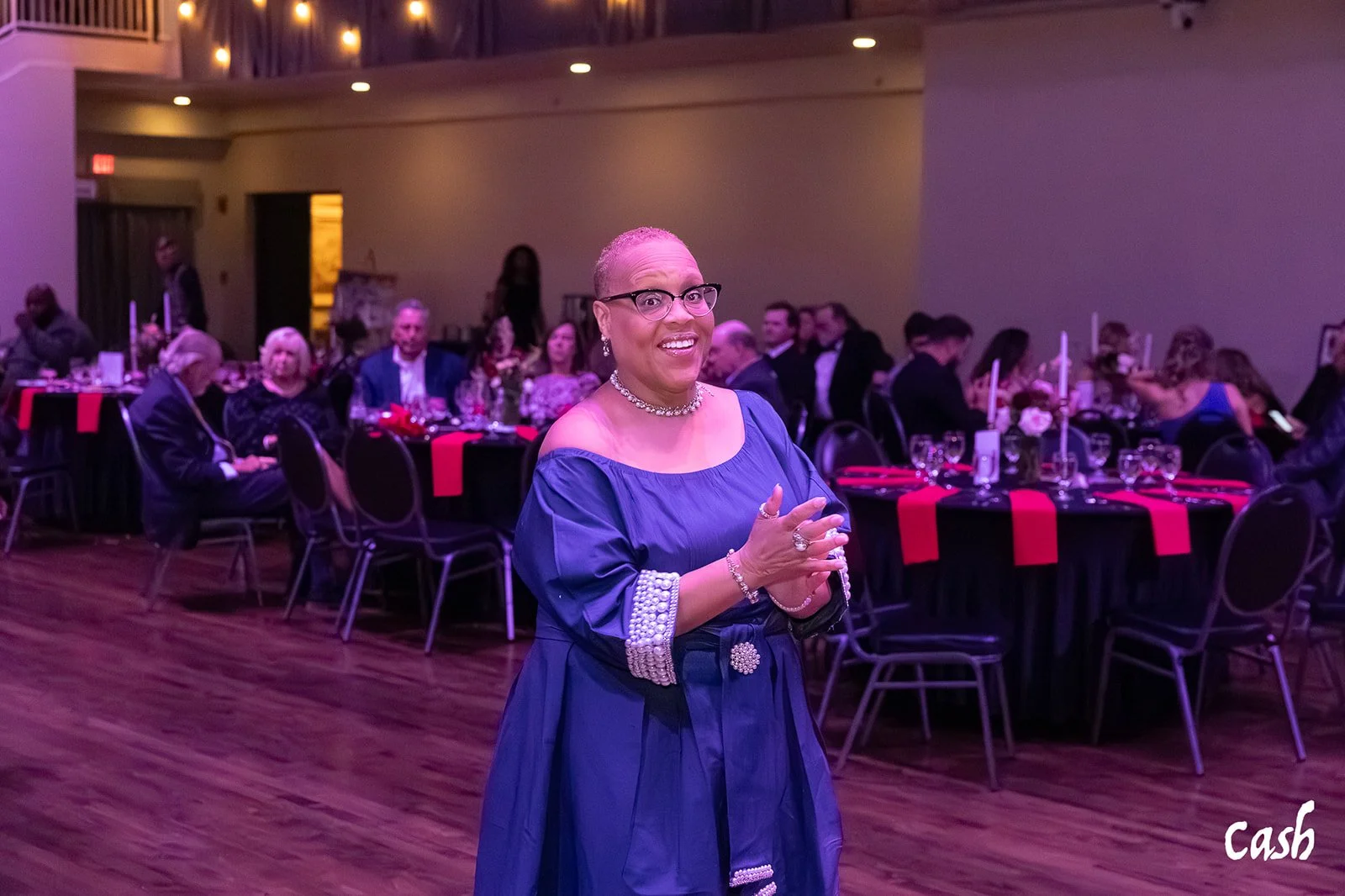 A woman in a royal blue dress with pearl accents, wearing glasses and jewelry, standing in a decorated event hall with tables set for a formal dinner, smiling at the camera.