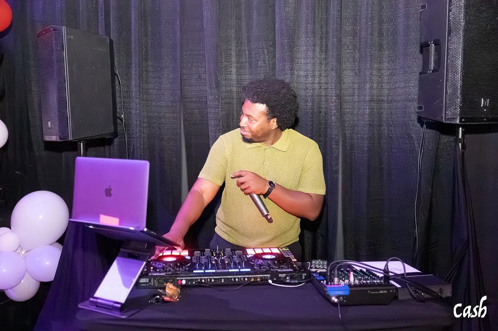 A DJ in a green short-sleeve shirt standing behind a mixing console at a DJ event, holding a microphone, with black curtains and speakers in the background, and white balloons on the left side.