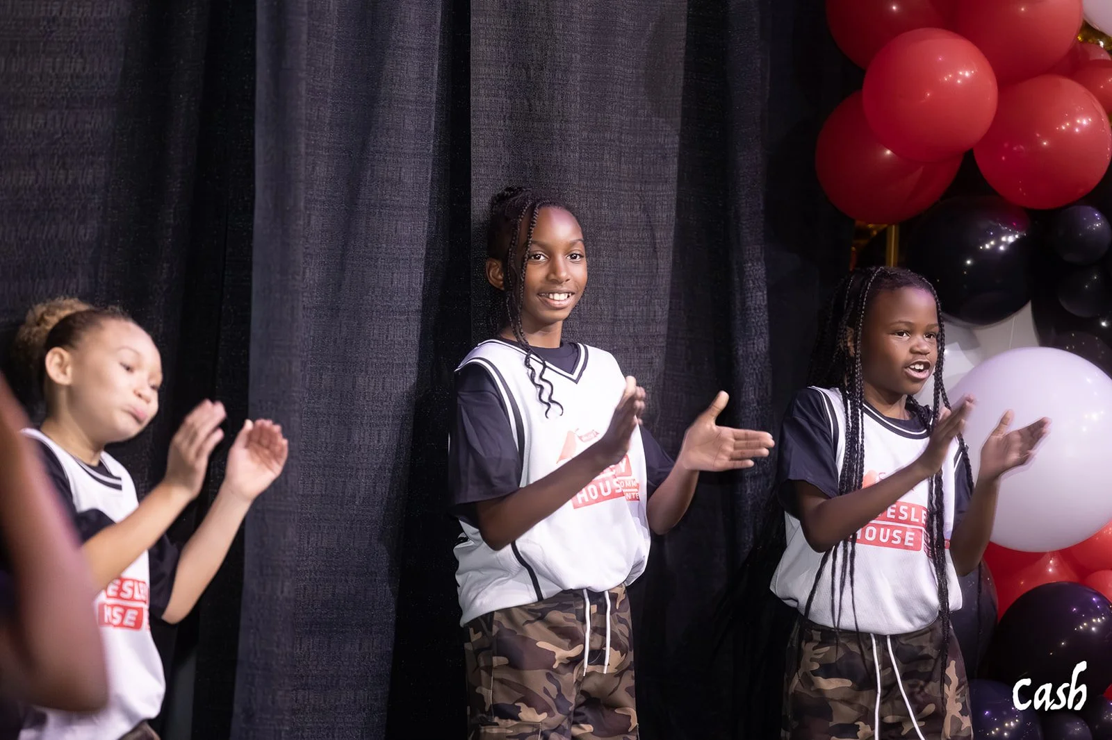Three young girls wearing matching sports jerseys, standing in front of black curtains with pink, black, and white balloons behind them, clapping and smiling.