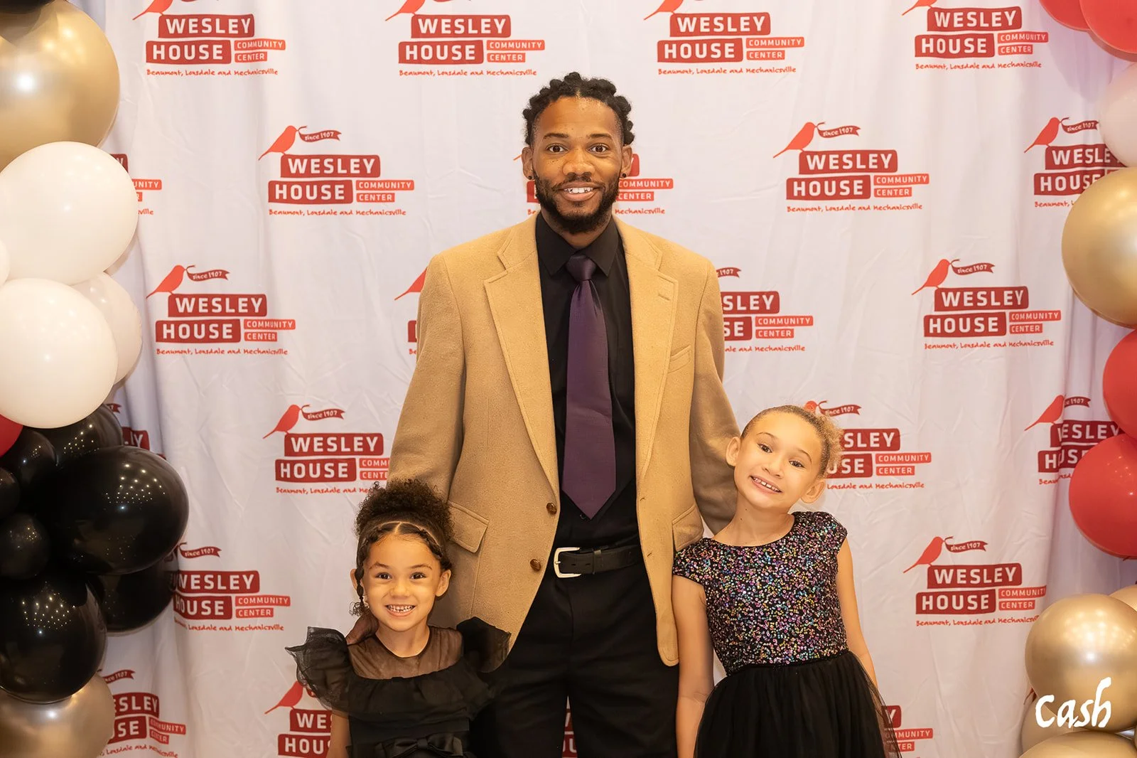 A man in a beige blazer and dark shirt with a purple tie poses with two young girls in colorful dresses, at an event with a backdrop featuring the Wesley House Community Center logo.
