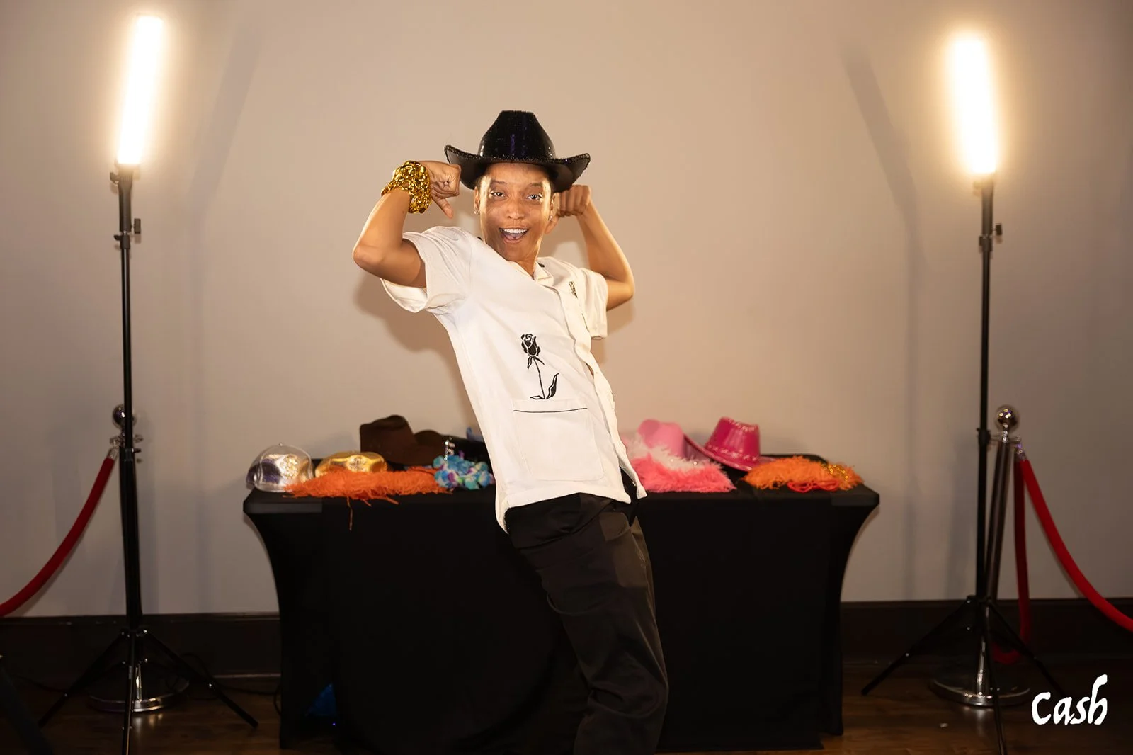 A person wearing a white shirt, black cowboy hat, and gold bracelet, smiling and posing with arms flexed in front of a display table with hats and accessories, in a room with two lights and a red rope barrier.