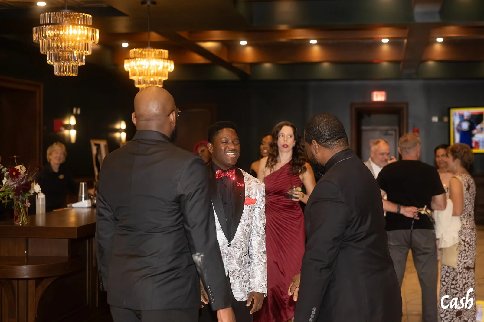 People socializing at a formal event in a dimly lit room with chandeliers, including a man in a silver tuxedo and others in black tuxedos and evening dresses.