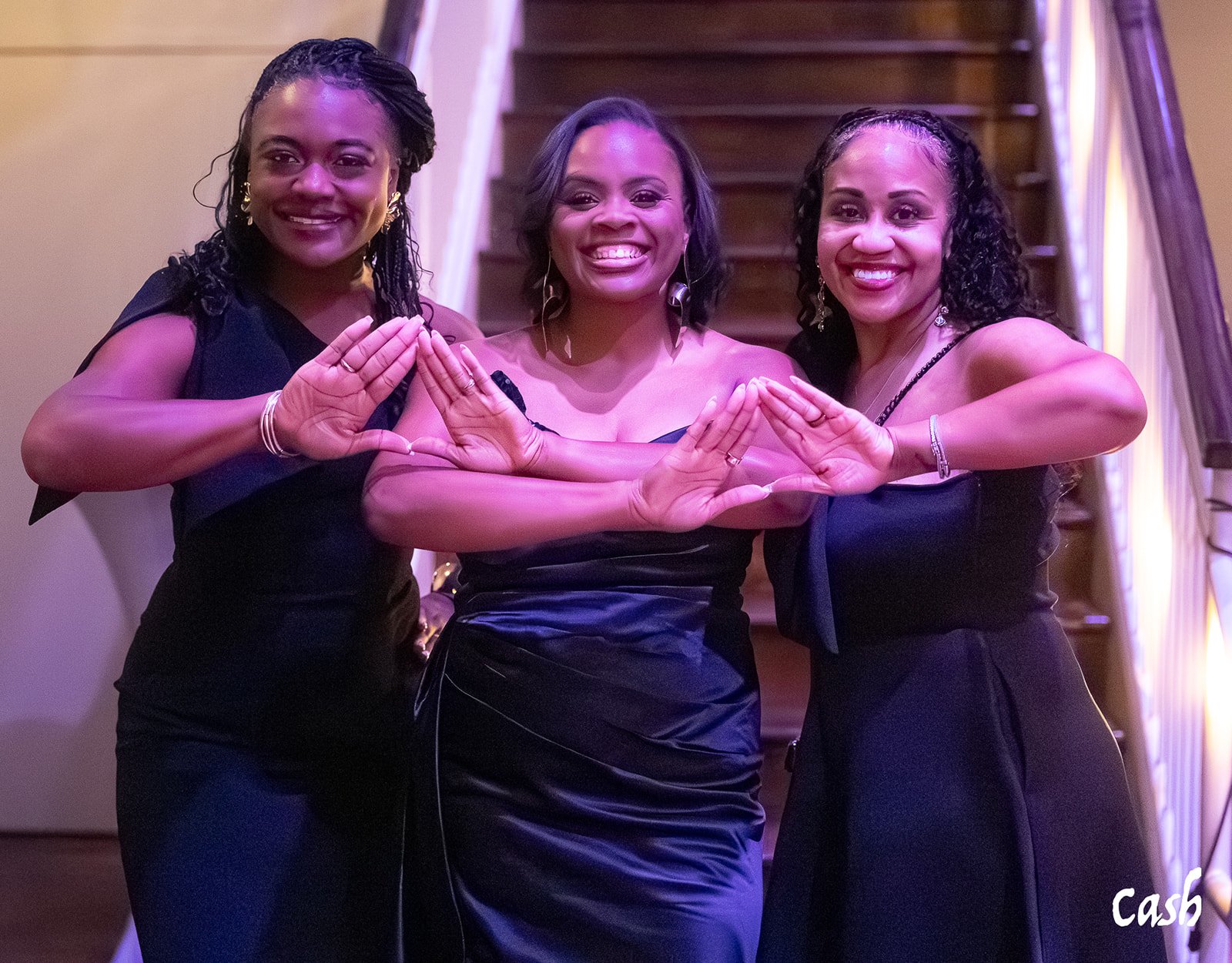 Three women wearing black dresses standing in front of a staircase, smiling and holding their hands together to form a triangle shape.