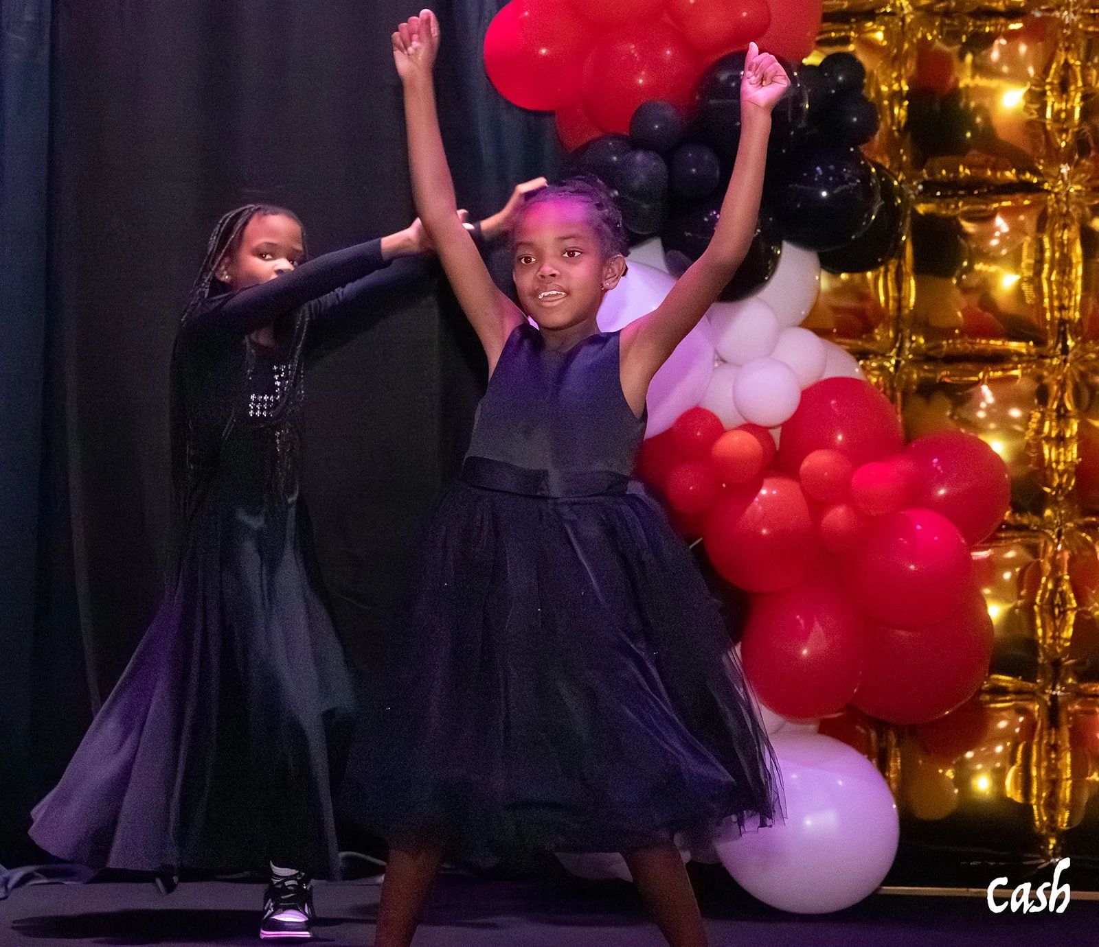 Two young girls dance in front of a balloon arrangement with black, red, white, and gold balloons at an event. One girl has arms raised and the other appears to be reaching out.